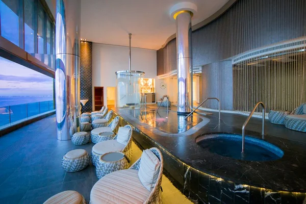 Luxurious spa with a hot tub, plush seating, and large windows offering a panoramic ocean view; modern and serene atmosphere.