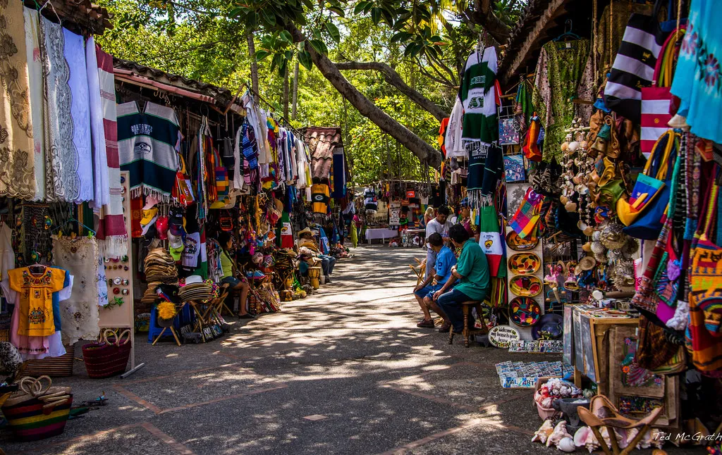 Lively outdoor market with colorful textiles, handicrafts, and people exploring stalls under lush trees.