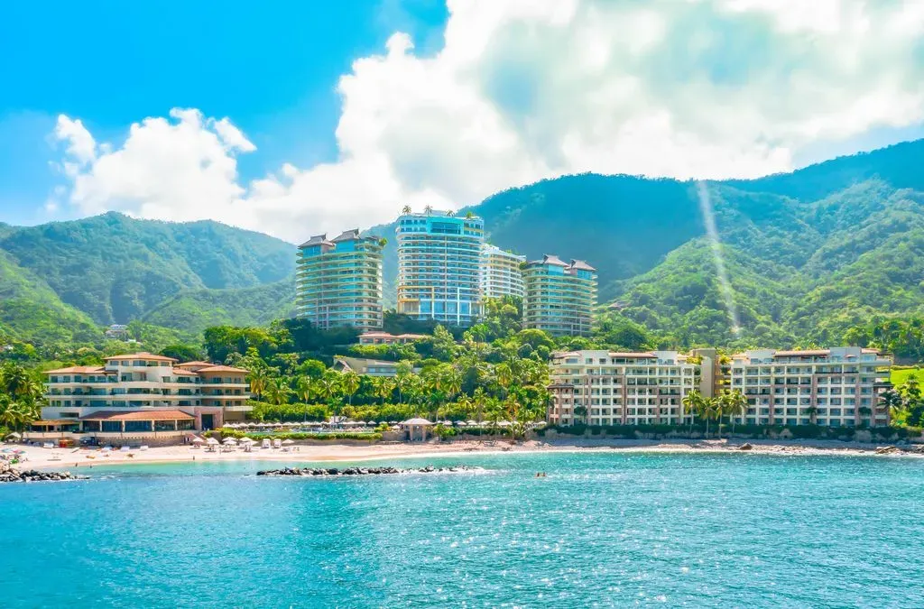 Aerial view of luxury beachfront resort with verdant mountains, pristine beach, and turquoise ocean under a bright sky.