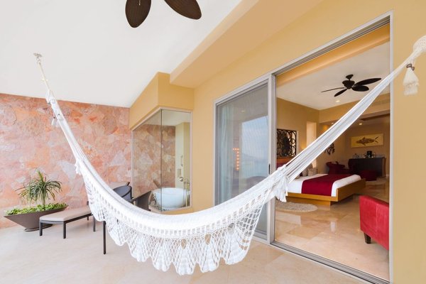 A white hammock hangs on a balcony with a view into a modern bedroom. The space features a stone wall, potted plant, and sliding glass doors.