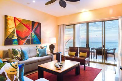 Modern living room with colorful abstract art, sofas, and a coffee table. Glass doors open to a balcony with an ocean view at sunset.