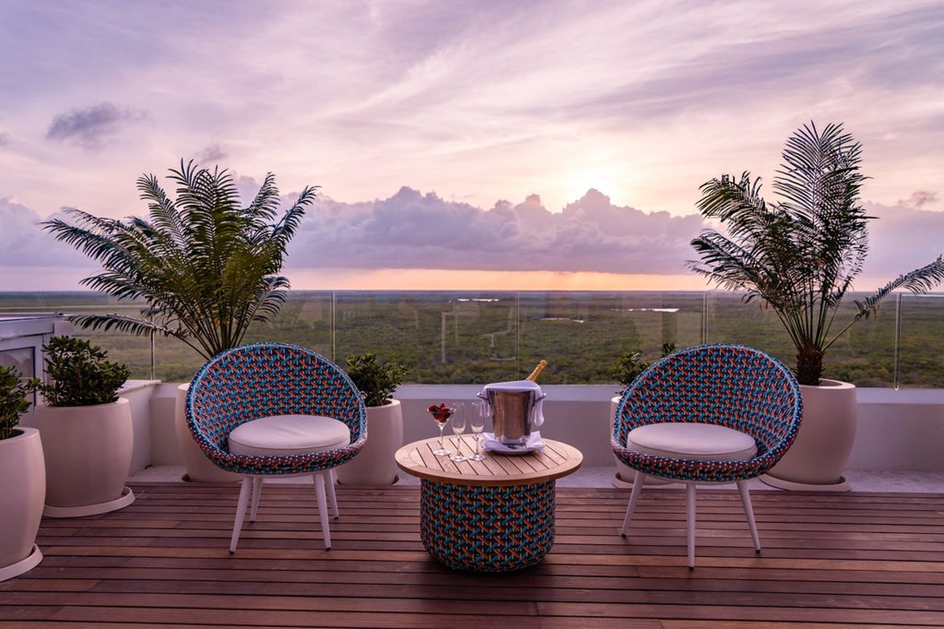 Rooftop patio with two patterned chairs, a small table with a champagne bucket, potted plants, and a scenic sunset view over a lush landscape.