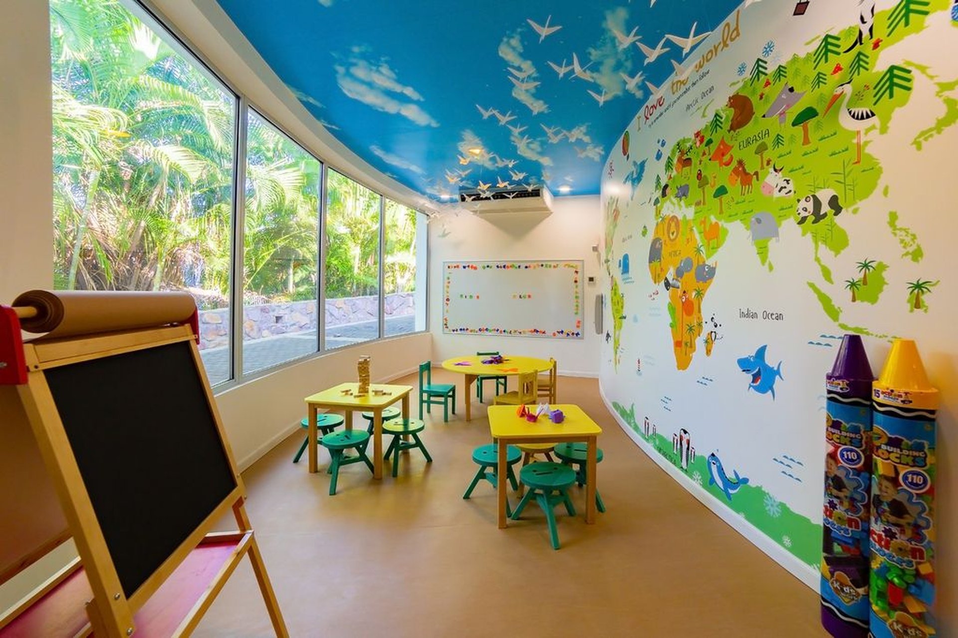 Colorful classroom with world map wall, small tables, chairs, and a chalkboard. Large windows and airplane ceiling decor enhance the bright space.
