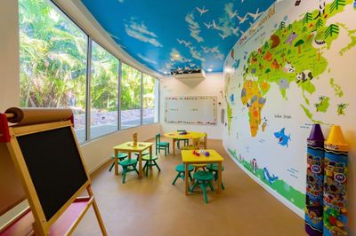 Colorful classroom with world map wall, small tables, chairs, and a chalkboard. Large windows and airplane ceiling decor enhance the bright space.