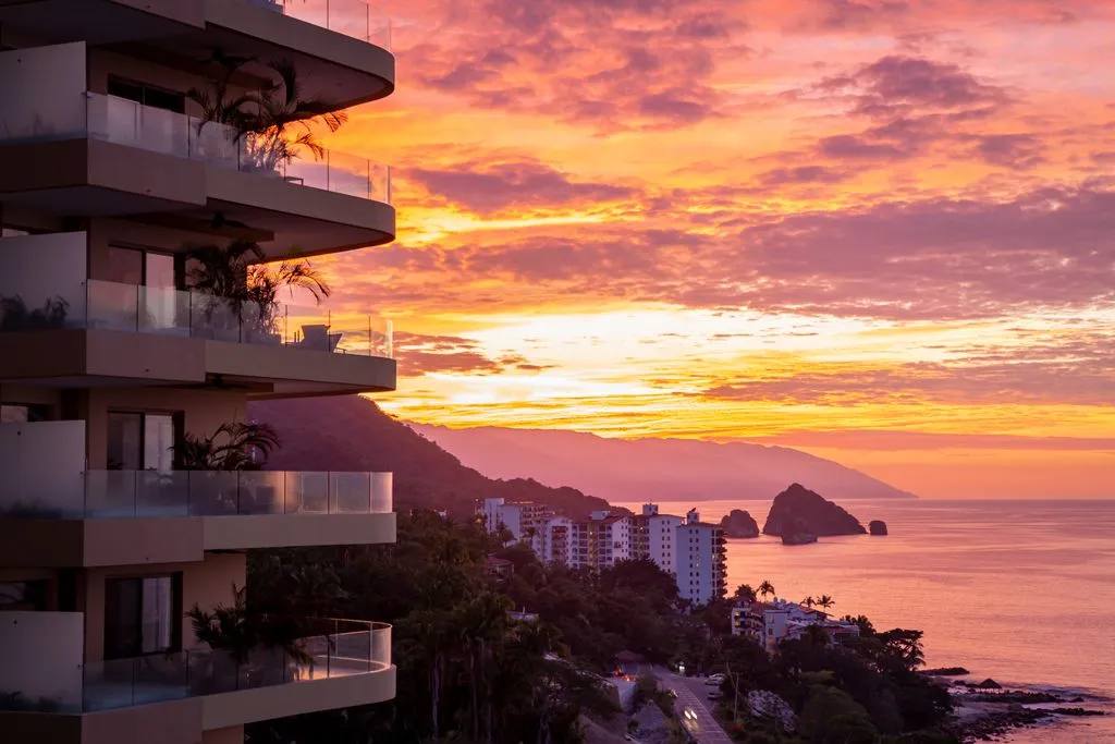 Sunset over a coastal city with a colorful sky, silhouetted mountains, a modern building in the foreground, and the ocean in the background.