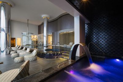 Luxurious spa interior with lounge chairs, a hot tub, cascading water feature, and ambient lighting creating a relaxing atmosphere.