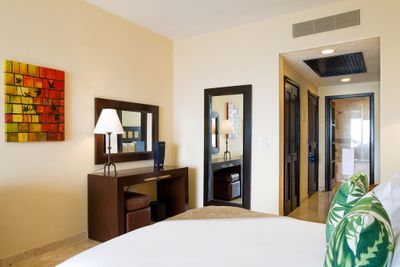 Hotel room with a bed, tropical-themed pillows, a wooden desk with a mirror and lamp, and a colorful wall art piece. Doorway leads to a bathroom.