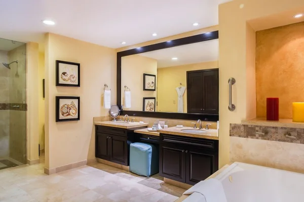 Modern bathroom with dual sinks, large mirror, and beige tiles. Art on walls, colorful candles by the bathtub, and recessed lighting.