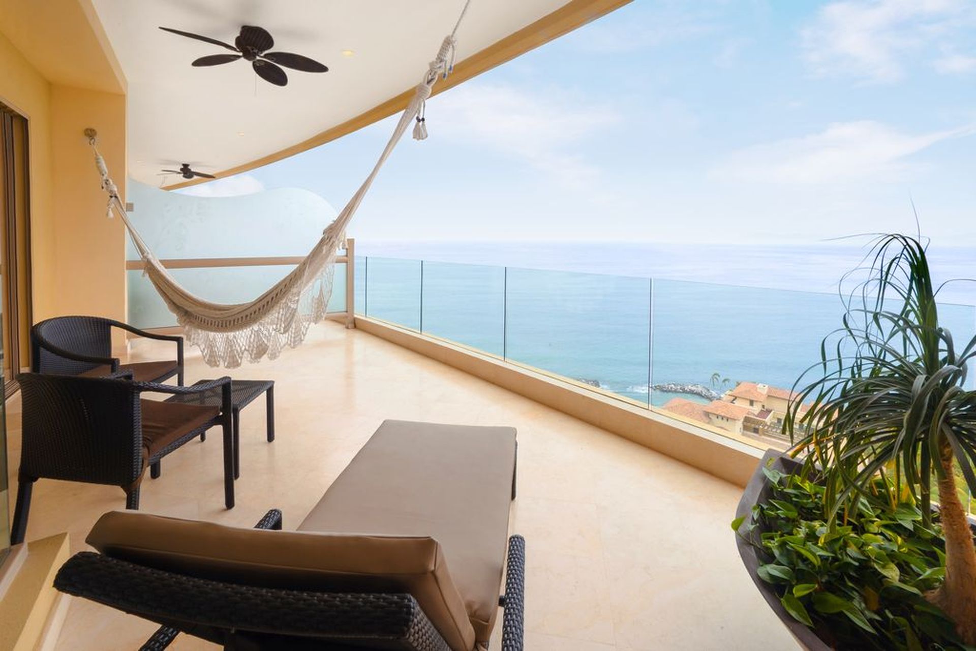 Spacious balcony with hammock, lounge chairs, and potted plant, overlooking a serene ocean view under a clear sky.