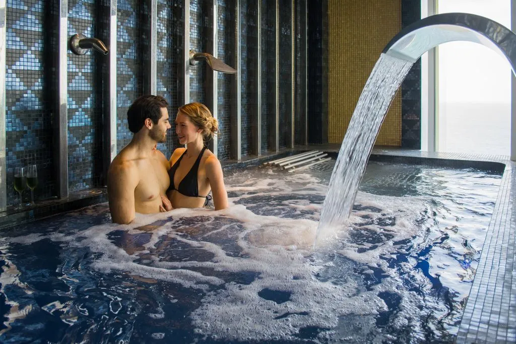 A couple enjoys a luxurious indoor whirlpool with cascading water, surrounded by elegant dark tiles.