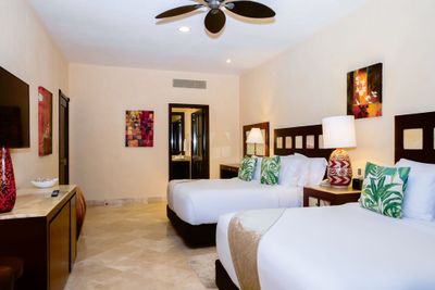 Hotel room with two double beds, tropical-themed pillows, modern decor, a ceiling fan, and warm lighting. Art adorns the walls.
