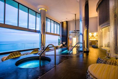 Luxurious indoor spa with hot tubs, elegant seating, and large windows offering a stunning ocean view at sunset.