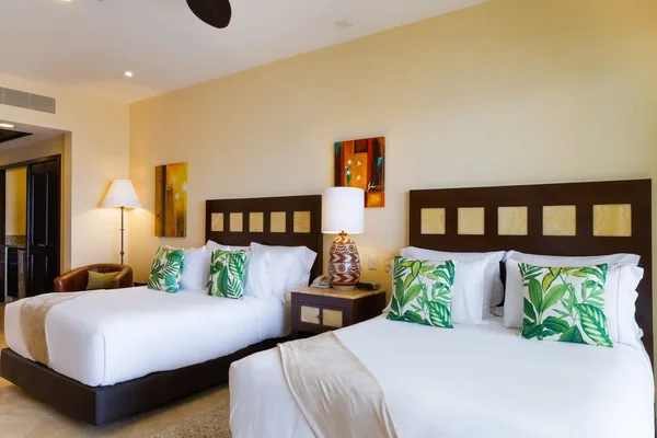 A hotel room with two double beds, each adorned with green leaf-patterned pillows, a lamp on a nightstand, and colorful wall art.