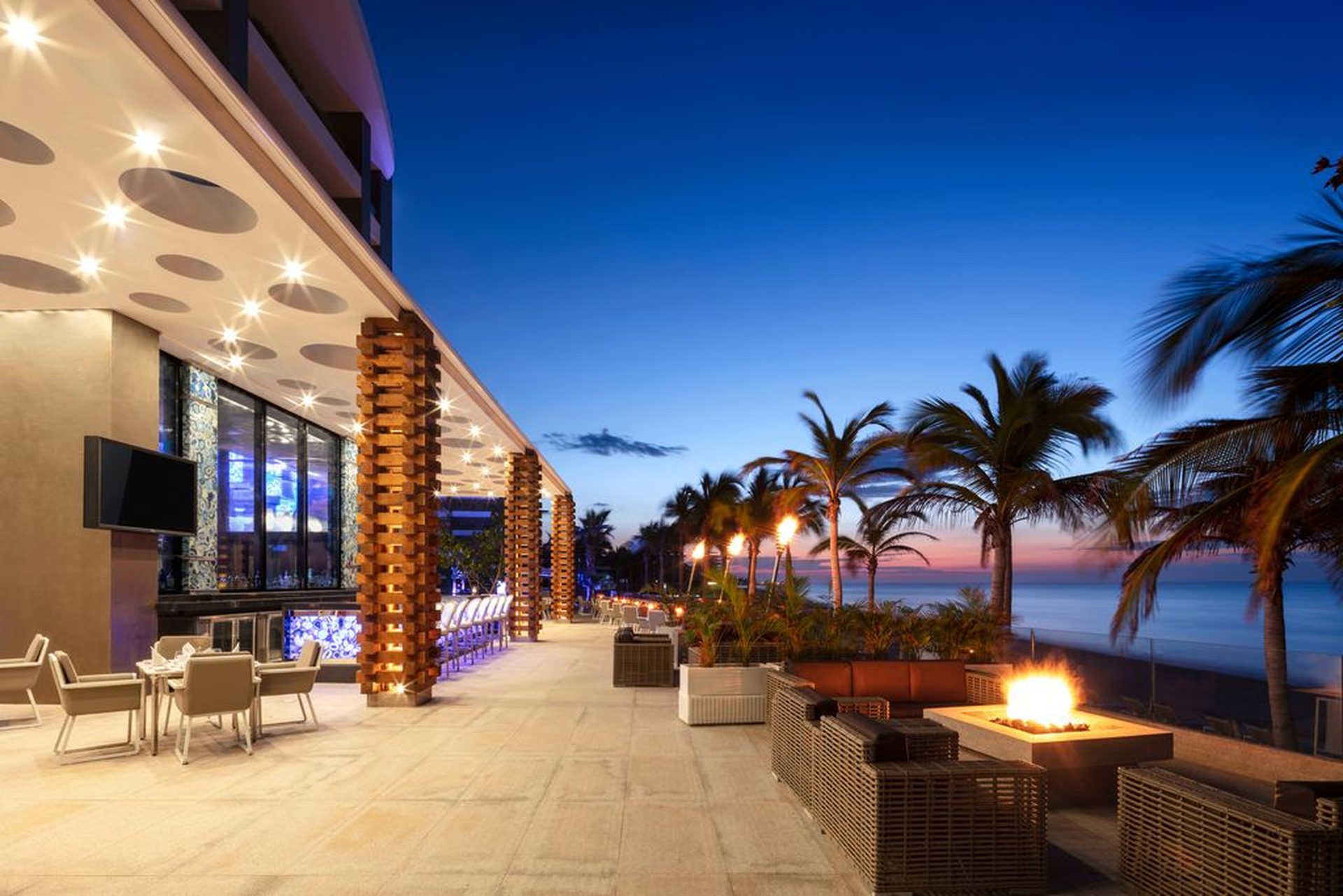Outdoor lounge with modern seating, fire pits, and palm trees overlooking the ocean at sunset, under a clear, deep blue sky.
