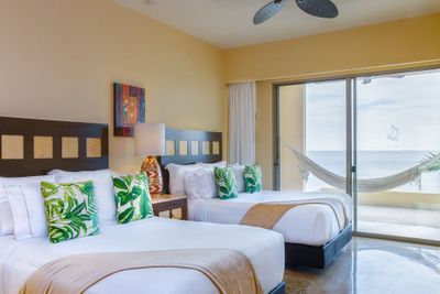 Bright hotel room with two double beds, tropical-themed pillows, a lamp, and a sliding glass door opening to a balcony with a hammock and ocean view.