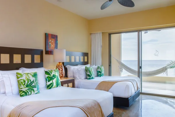 Bright hotel room with two double beds, tropical-themed pillows, a lamp, and a sliding glass door opening to a balcony with a hammock and ocean view.