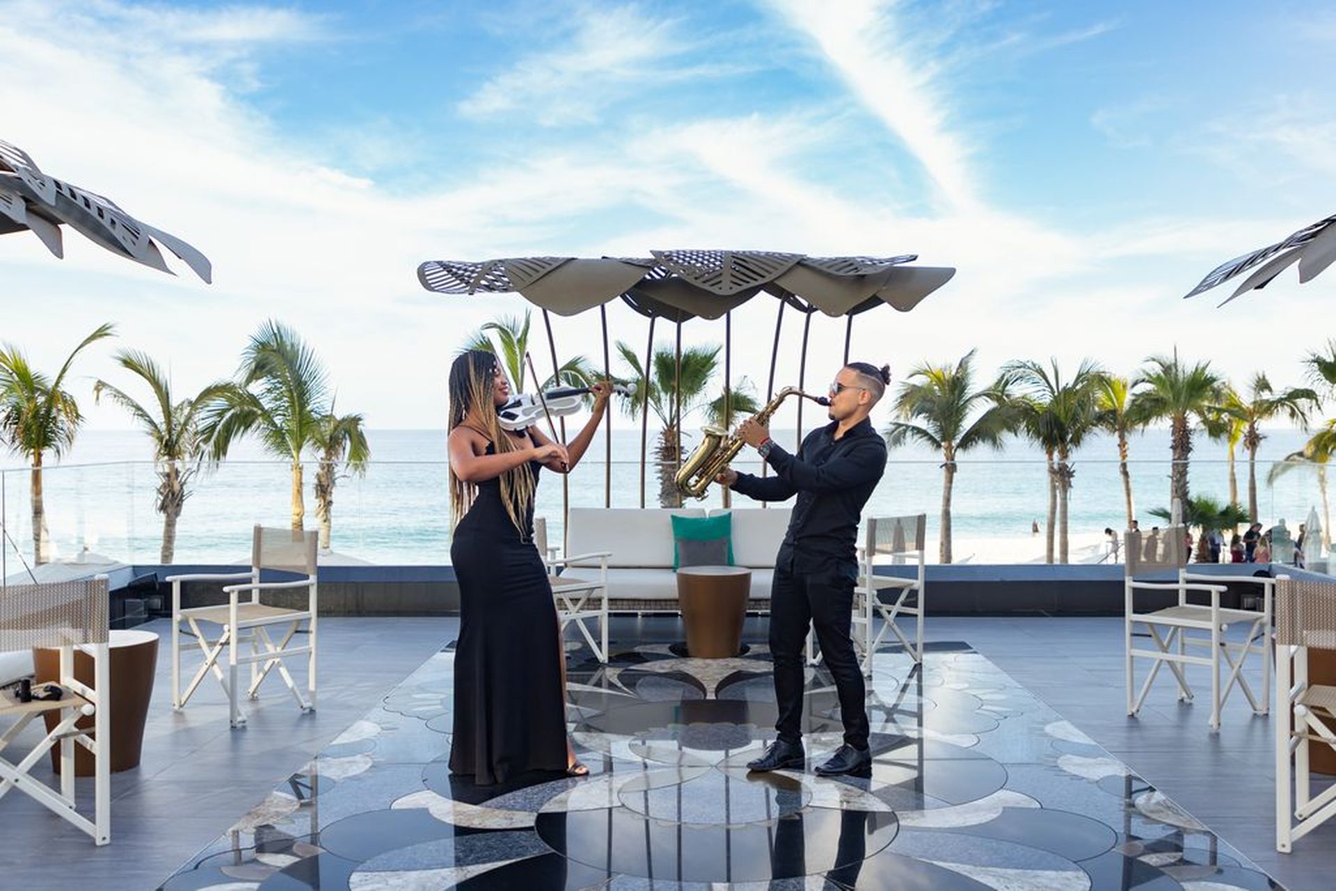 A violinist and saxophonist perform on a stylish outdoor patio by the ocean, surrounded by palm trees and modern decor.