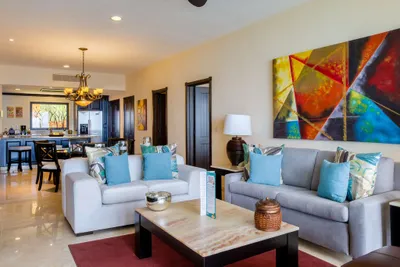 Modern living room with two sofas, colorful abstract art, and a marble coffee table. Dining area and kitchen visible in the background.