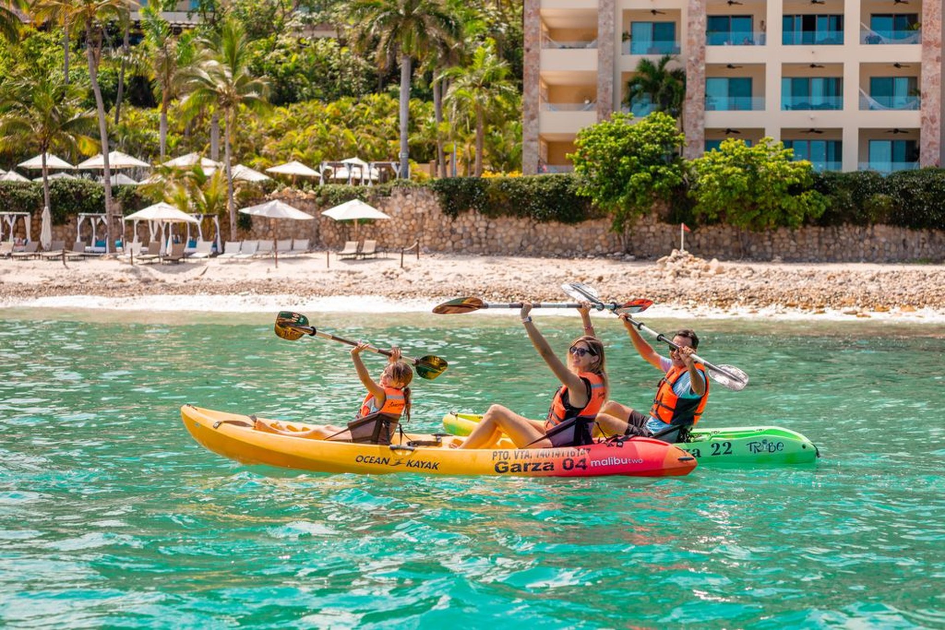 A family kayaking in turquoise waters near Garza Blanca Cancun resort, bordered by a sandy beach and lush palm trees in the background.