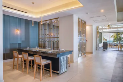 Modern dining area with a sleek black table, wooden chairs, soft lighting, and decorative metal partitions, overlooking a view of palm trees.