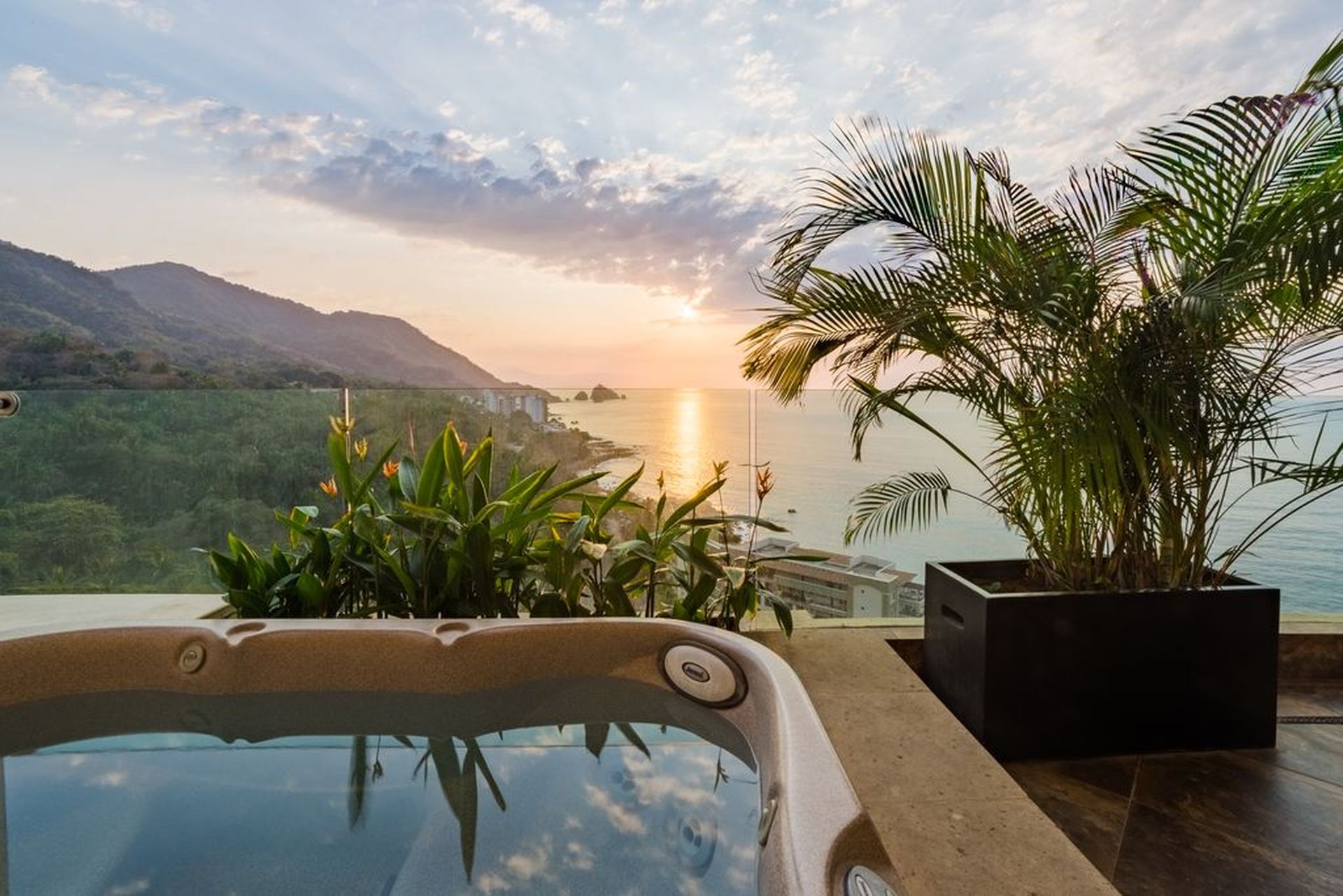 A luxurious hot tub overlooks a scenic ocean view at sunset, framed by lush tropical plants and distant mountains.