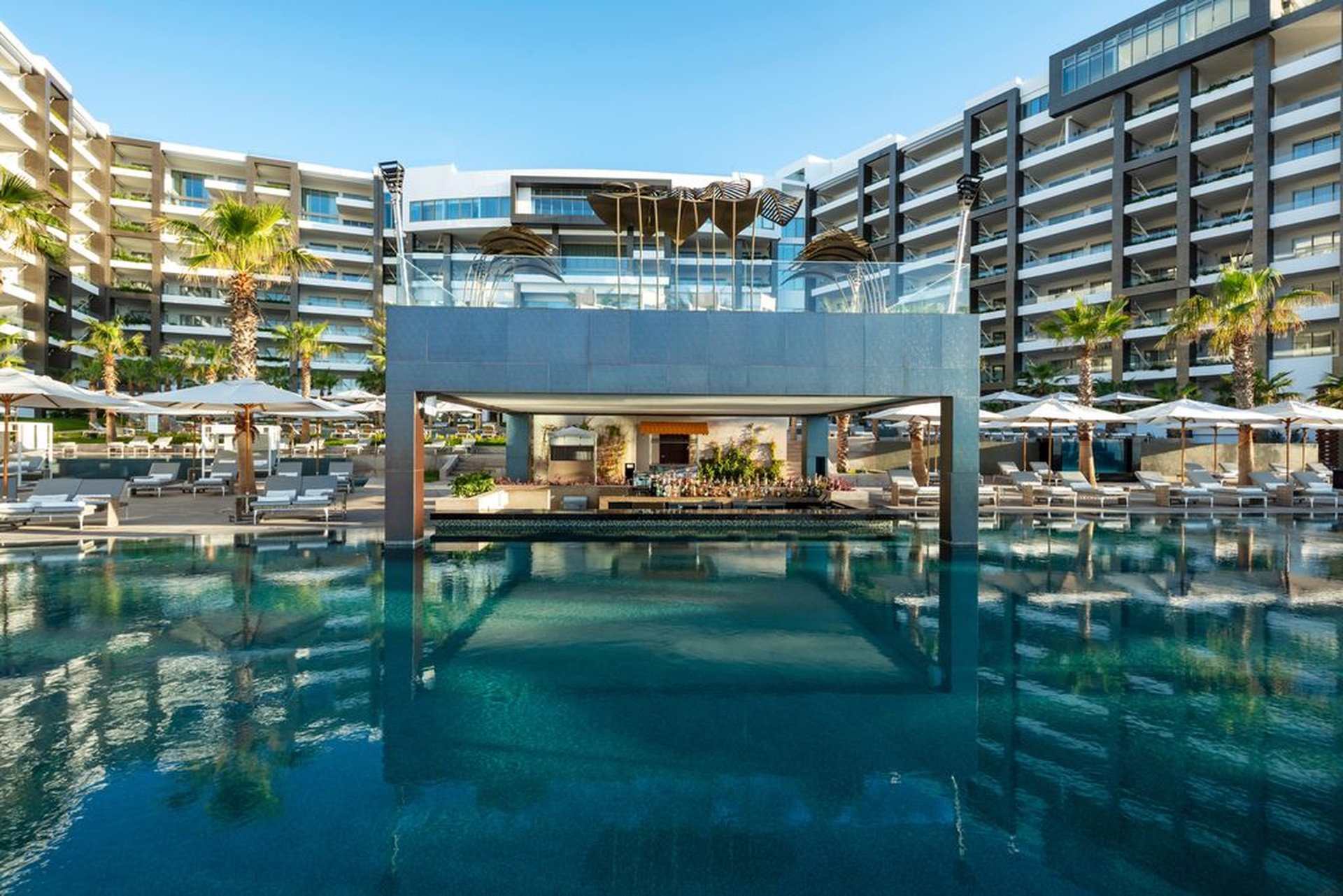Luxurious hotel pool area with modern architecture, surrounded by palm trees and sun loungers, reflecting in the calm water under a clear sky.
