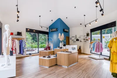 Modern boutique interior with wooden floors, clothing racks, mannequins, and a blue accent wall. Large windows provide natural light.