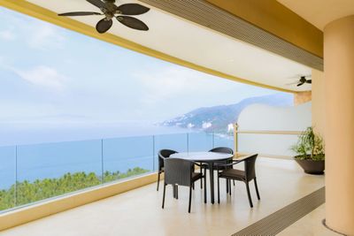 Spacious balcony with a dining set, ceiling fans, and glass railing overlooking a scenic ocean view and distant mountains.