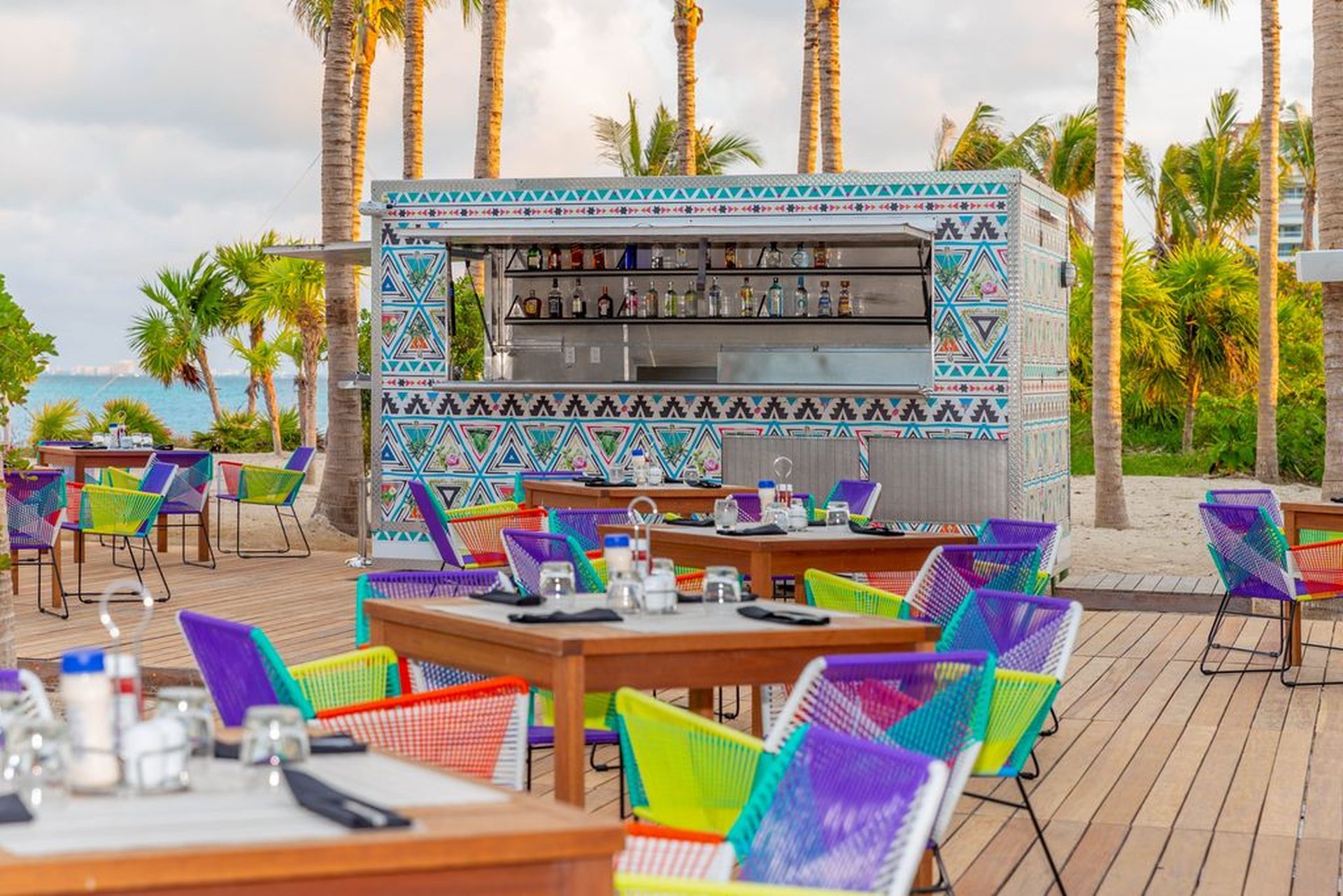 Colorful outdoor beach bar with vibrant chairs, wooden tables, and palm trees in the background under a partly cloudy sky.