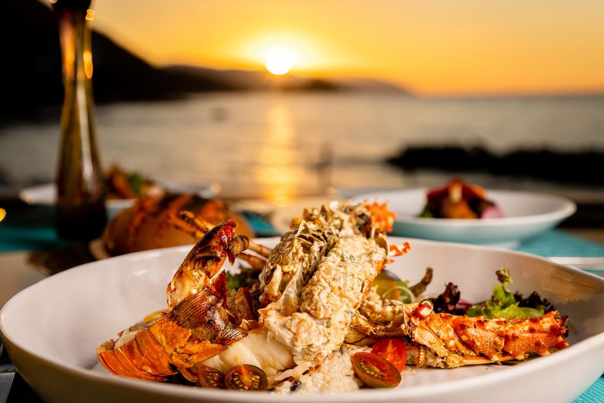A plate of lobster with salad and cherry tomatoes is served at an outdoor dining table, with a sunset over the ocean in the background.