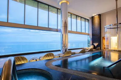 Luxurious indoor pool area with wicker chairs, large windows offering a panoramic ocean view, and modern decor with soft lighting.