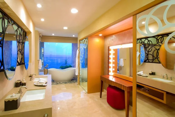 Luxurious bathroom with a large mirror, double sinks, a vanity with a red stool, a bathtub, and a view of the ocean through large windows.