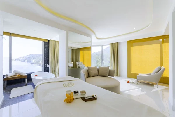 Luxurious spa room with a massage table, bathtub, and large windows offering a scenic ocean view. Soft lighting and elegant decor create a serene ambiance.