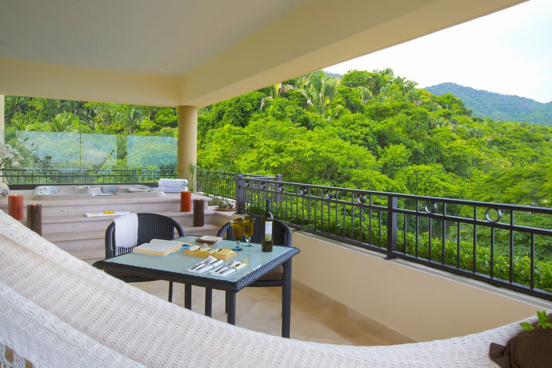Covered patio with a hammock, table set for two with wine, overlooking lush green forest and hills. Relaxing outdoor retreat.