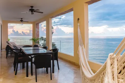 Covered terrace with dining table, hammock, and ceiling fans overlooking the ocean at sunset, framed by large open windows.