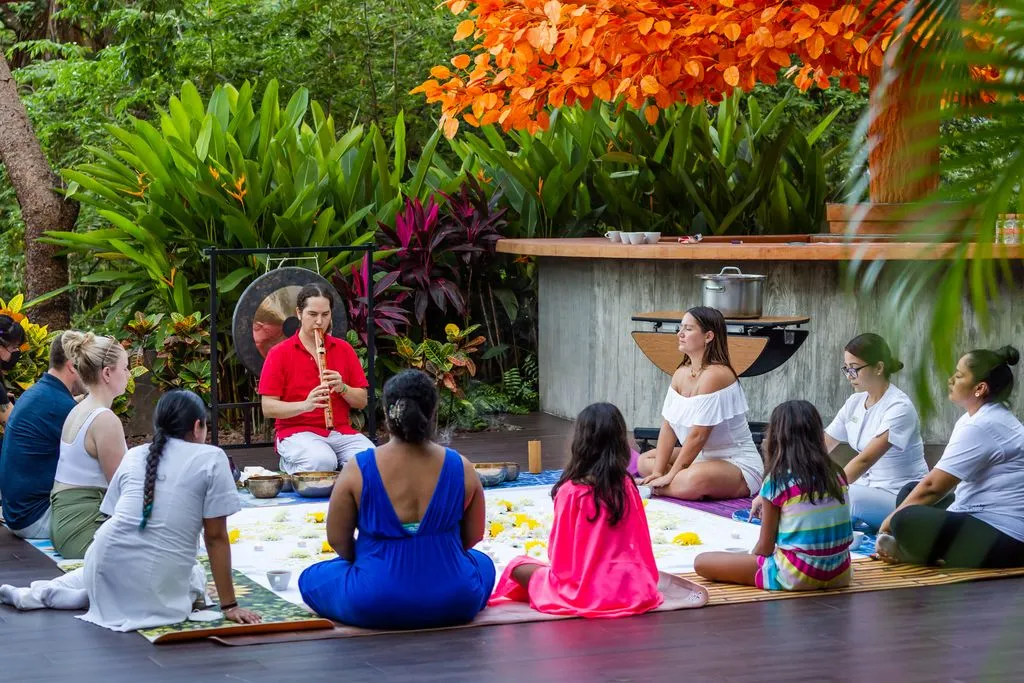 A group sits in a circle outdoors, listening to a musician playing a flute, surrounded by lush greenery and vibrant orange foliage.