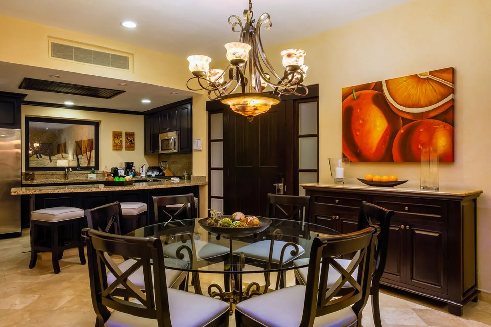 Elegant kitchen with dark wood cabinets, a glass dining table, cushioned chairs, a decorative chandelier, and vibrant fruit-themed paintings.