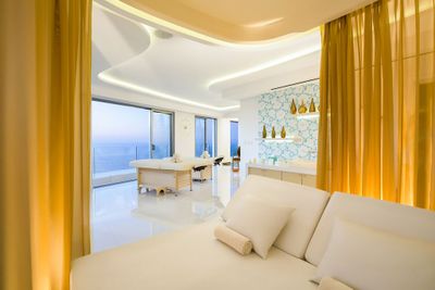 Luxurious spa room with white furniture, golden curtains, massage tables, and a view of the ocean through large windows.