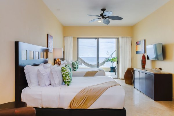 Bright hotel room with two beds, tropical decor, a ceiling fan, and a view of the ocean through sliding glass doors. A hammock is visible outside.
