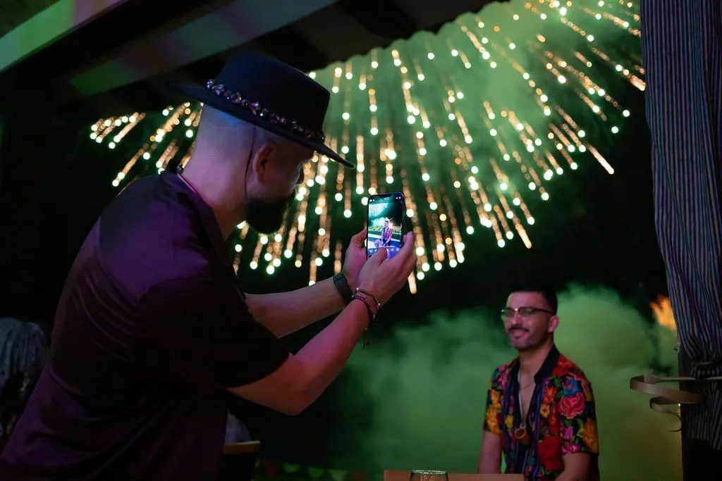 Man photographs another man with fireworks in the background, capturing a festive moment at night.