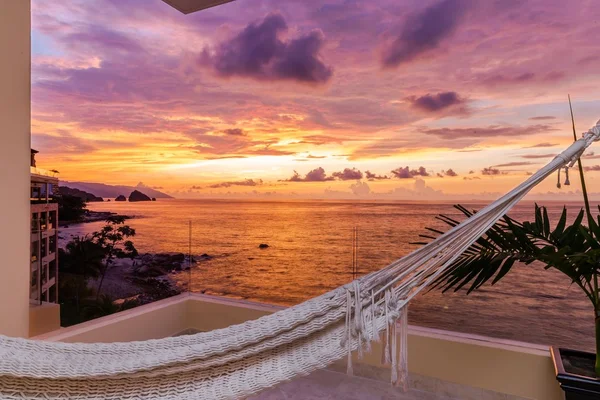Hammock on a balcony overlooking a vibrant ocean sunset with pink and orange hues, palm leaves, and distant rocks near the shore.