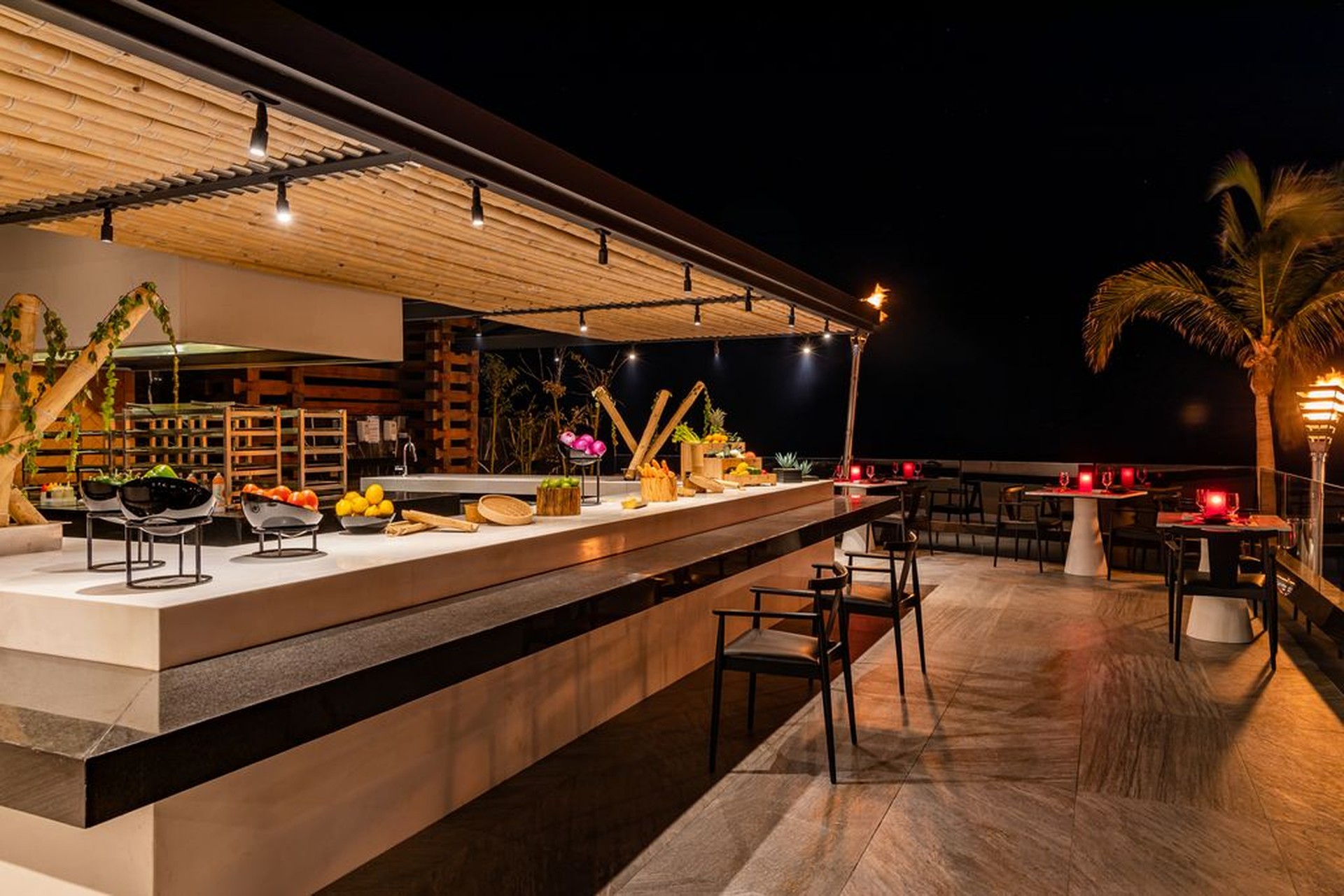 Outdoor restaurant bar at night with modern decor, wooden ceiling, tropical plants, and ambient lighting. Tables set with red candles.