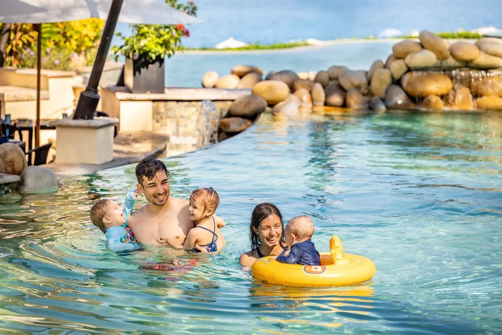 A family enjoys swimming in a luxurious resort pool with a scenic ocean view, surrounded by rocks and tropical plants.