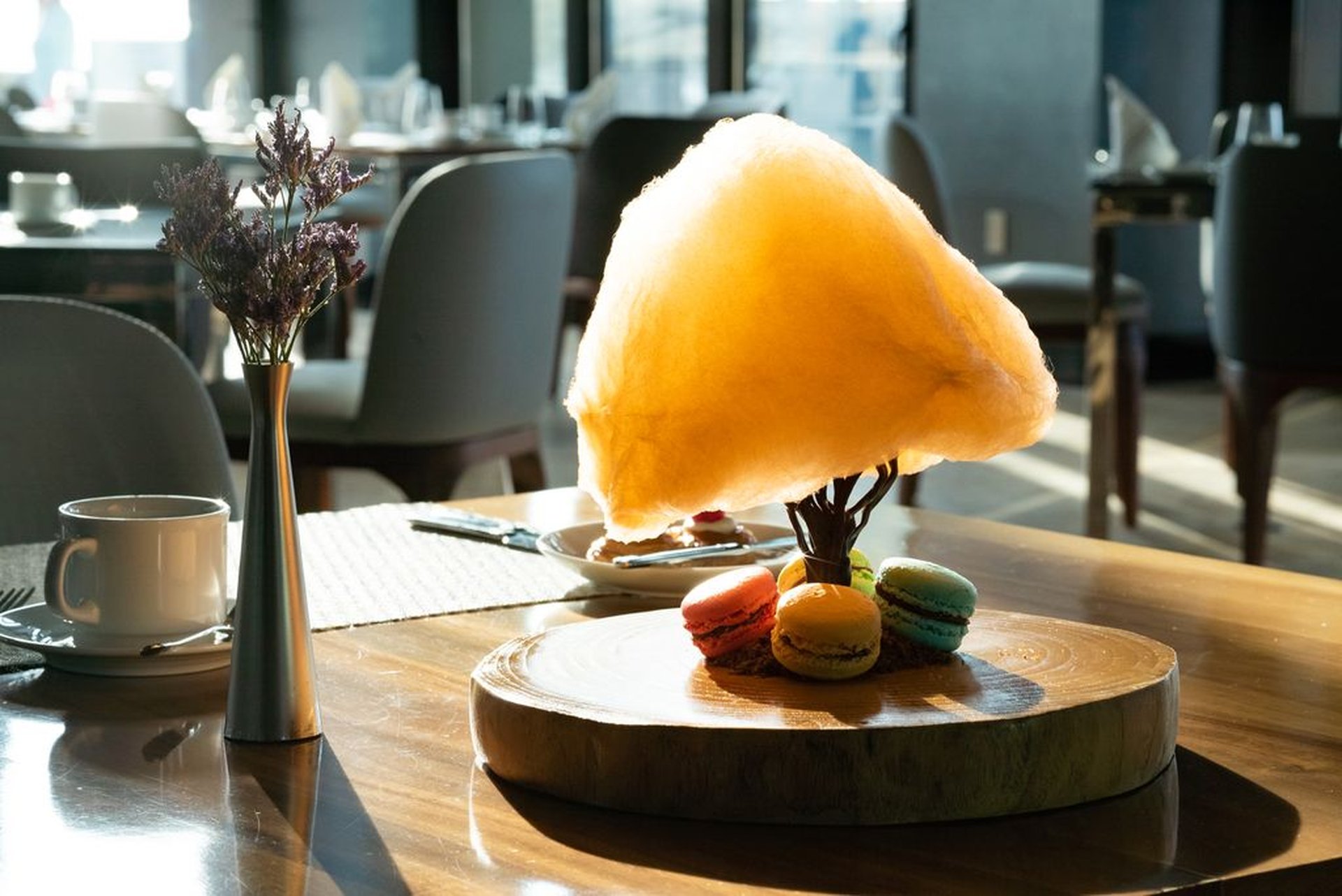A wooden platter with colorful macarons and a large, fluffy cotton candy tree on a sunlit table in a modern dining setting.
