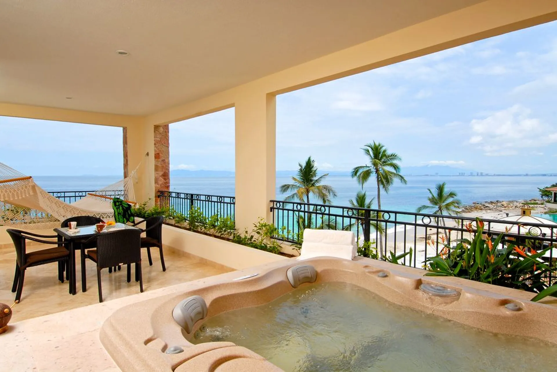 Outdoor balcony with a hot tub, hammock, dining table, and ocean view. Palm trees and a clear sky enhance the tropical setting.