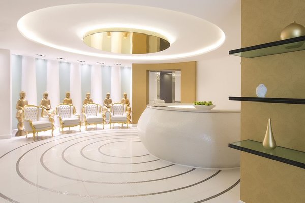 Modern lobby with a circular reception desk, gold accents, decorative vases, and a row of elegant chairs. Ceiling features a large round light.