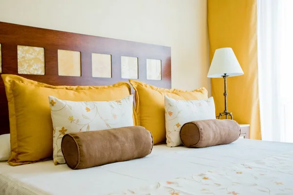 Cozy bedroom with a double bed, yellow pillows, floral cushions, brown bolster pillows, a wooden headboard, and a bedside lamp.