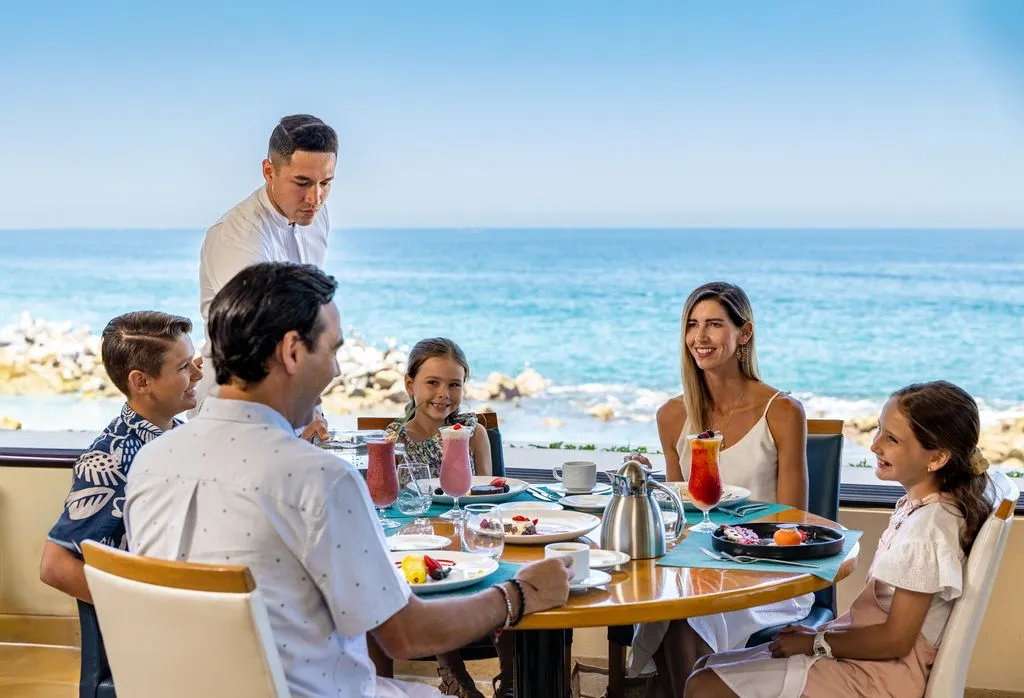 A family enjoys a gourmet breakfast by the ocean at Garza Blanca Cancun, with a waiter serving drinks and a view of the sparkling blue sea.