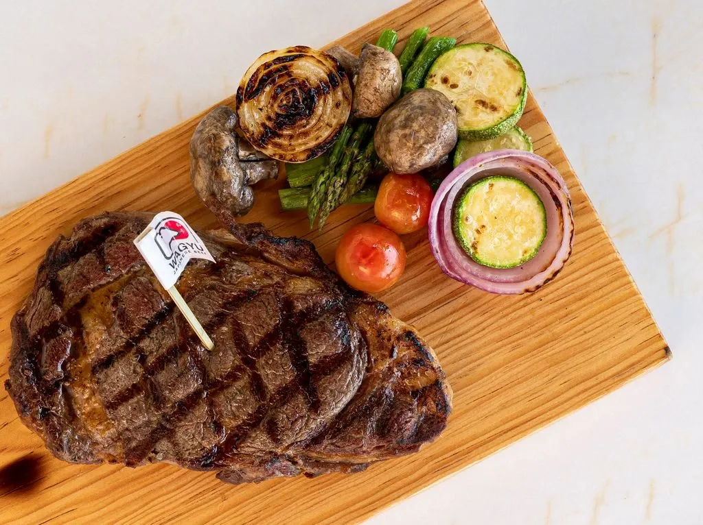 Grilled steak with a flag on a wooden board, served with grilled vegetables like mushrooms, zucchini, tomatoes, and onions.