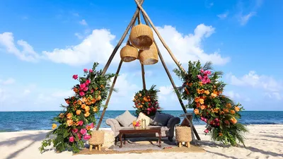 Luxurious beach setup with vibrant floral arrangements, elegant seating, and ocean backdrop under a clear blue sky.
